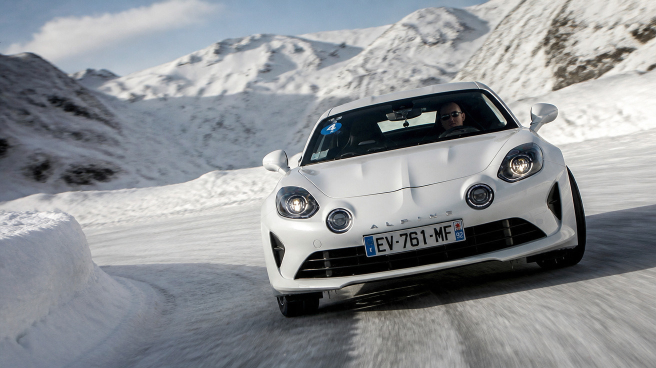 Carro esportivo branco em curva na estrada com céu azul e montanhas cobertas de neve ao fundo.