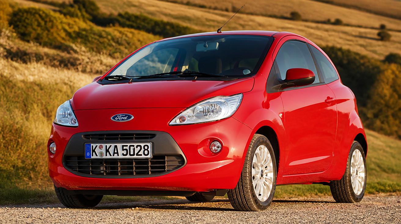 Carro Ford vermelho estacionado em estrada de terra com vegetação ao fundo em área rural.