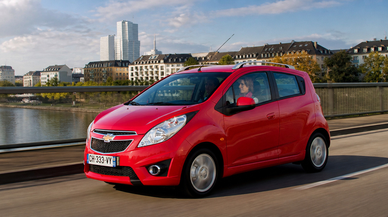 Carro vermelho Chevrolet em ponte com rio e prédios ao fundo durante o dia ensolarado.